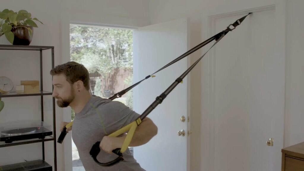 Man exercises with TRX straps anchored to a door at home.