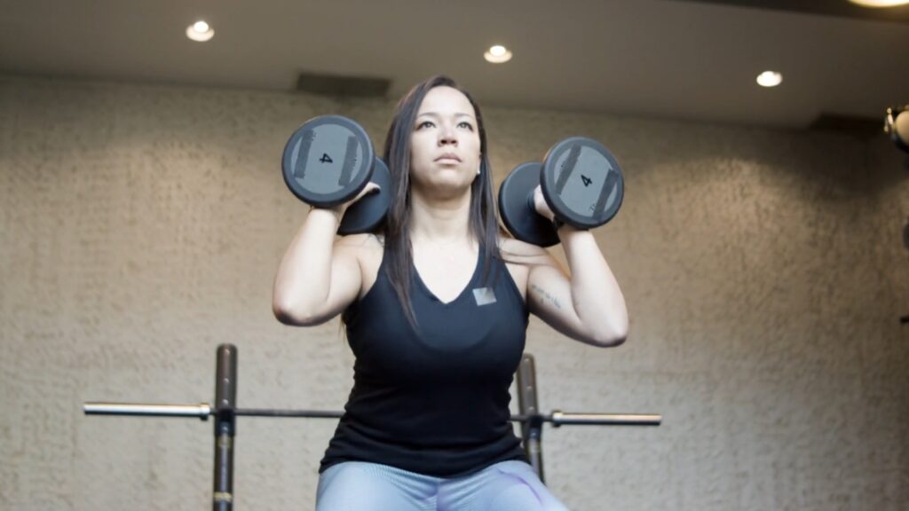 A woman holds two dumbbells at shoulder height during a gym workout.