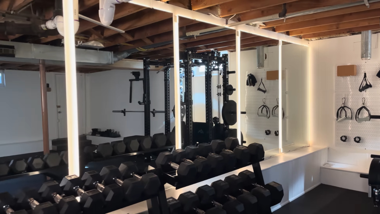 Basement gym with dumbbell racks, wall mirrors, LED lights, and exposed ceiling beams.