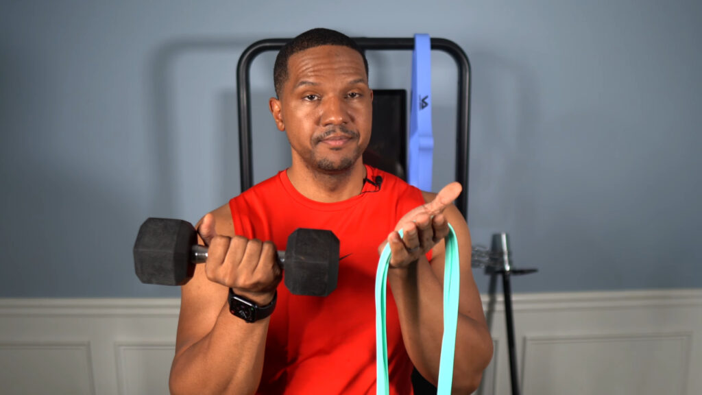 A man in a red shirt holds a dumbbell and a resistance band in his home gym.