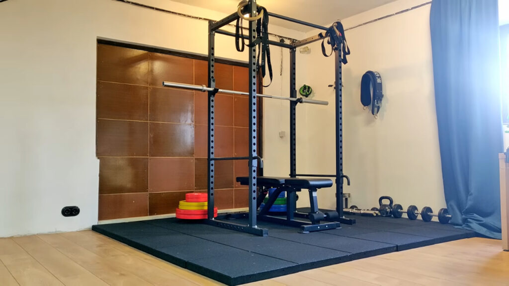 Apartment home gym with a squat rack, weight bench, dumbbells and rubber floor mats.