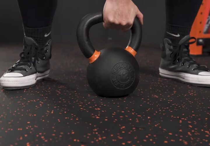A person holds a kettlebell above black rubber garage gym flooring with red speckles.
