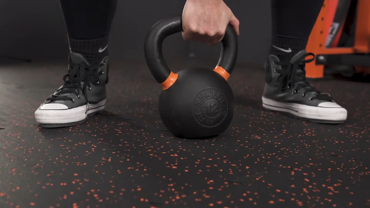 A person holds a kettlebell above black rubber garage gym flooring with red speckles.