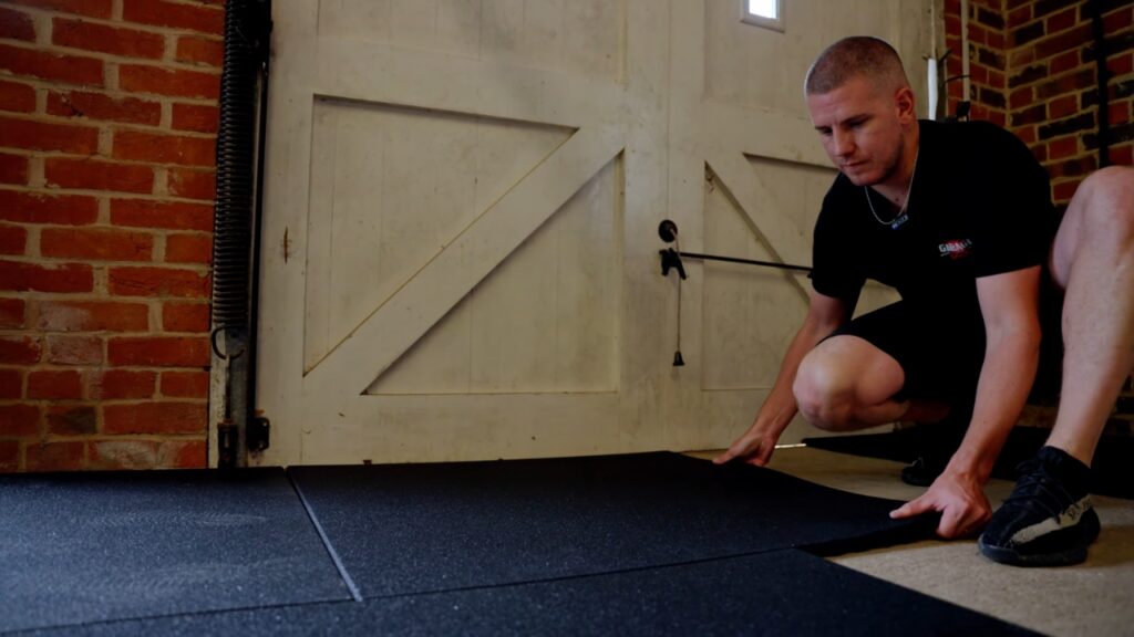 A man installs black rubber gym flooring near the garage door.