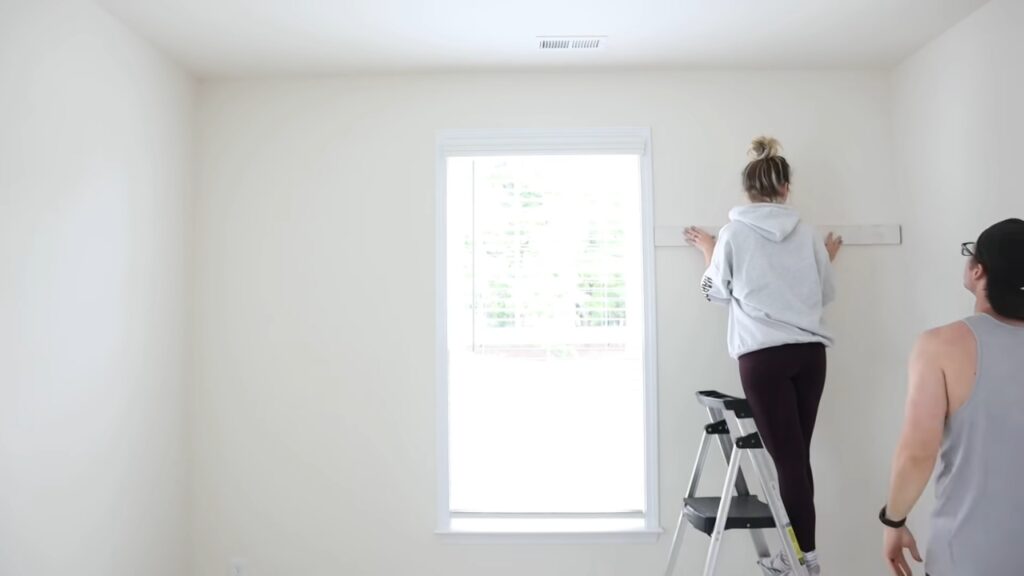 A woman stands on a stepladder to measure a wall as a man watches in an empty room.