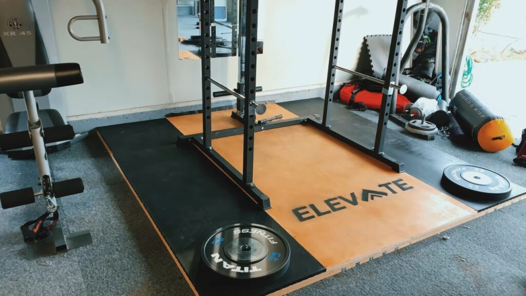 A garage gym lifting platform with a wooden center, black rubber mats and a squat rack.