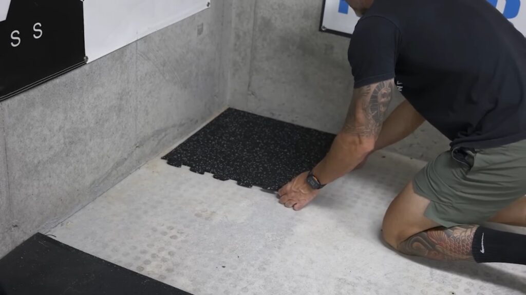 A man installs black interlocking rubber gym tiles on a concrete garage floor.