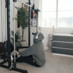 A person in a gray hoodie uses a cable machine in a carpeted apartment home gym.