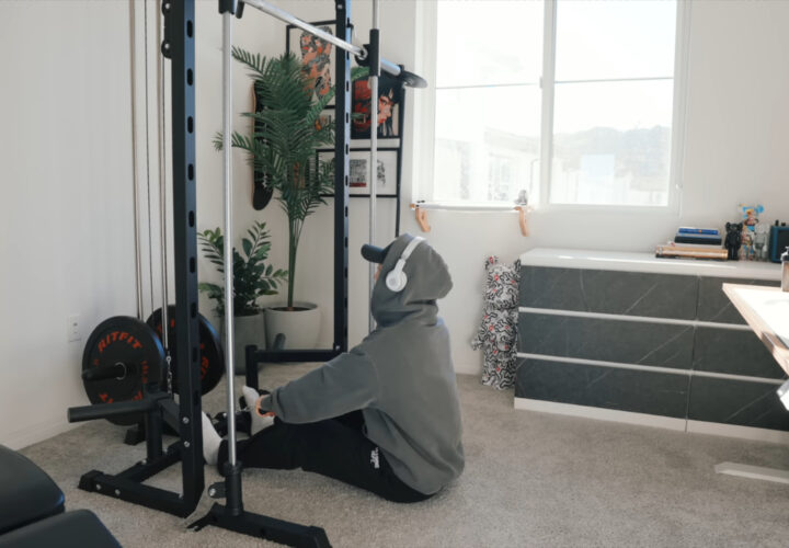 A person in a gray hoodie uses a cable machine in a carpeted apartment home gym.