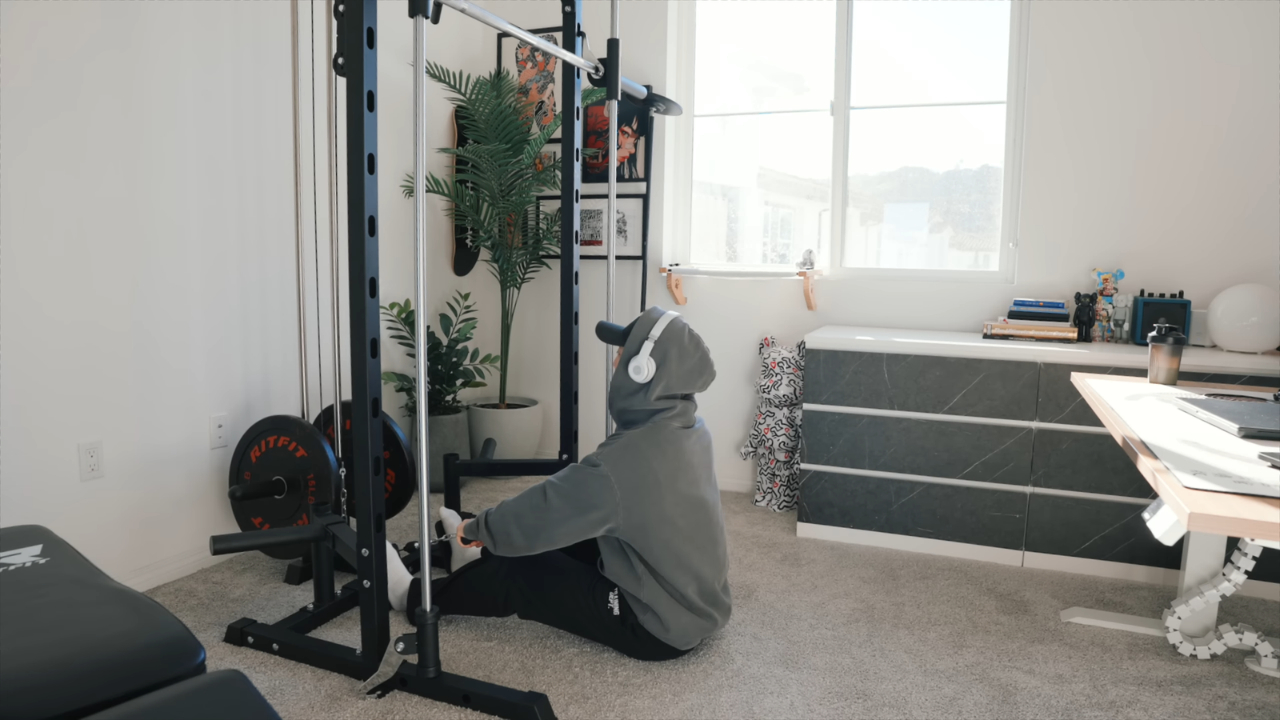 A person in a gray hoodie uses a cable machine in a carpeted apartment home gym.