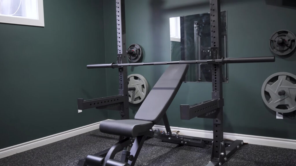 A small home gym with a wall mirror, weight bench, barbell rack, and weight plates.