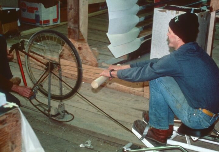 Two men test an early Concept2 rowing machine prototype made with a bicycle wheel inside a Vermont barn.