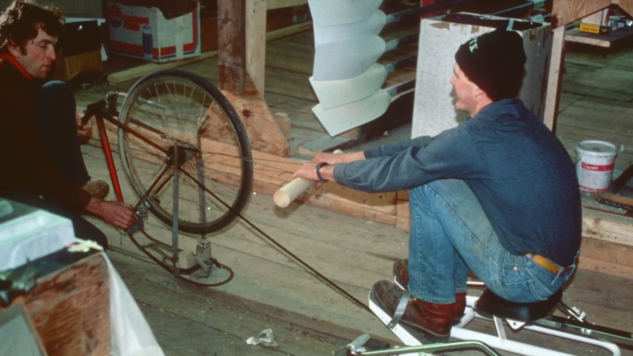 Two men test an early Concept2 rowing machine prototype made with a bicycle wheel inside a Vermont barn.