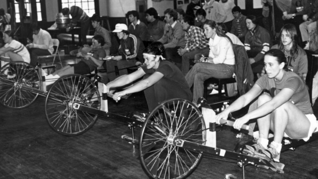 Rowers compete on early Concept2 rowing machines with large bicycle wheels as spectators watch.