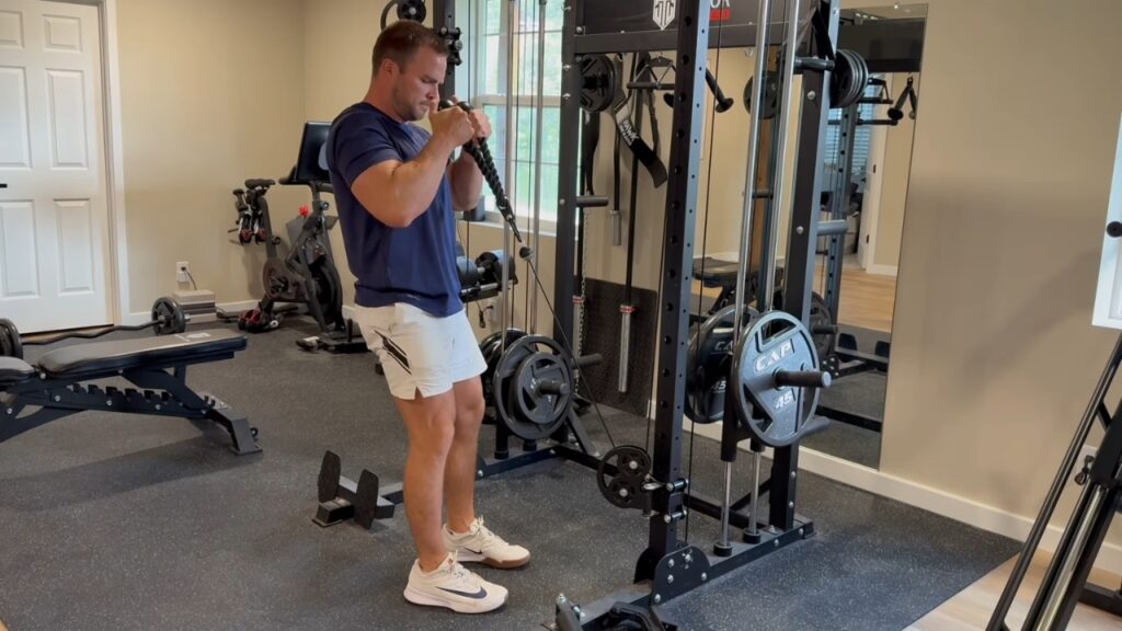 A man performs cable rope curls in his home gym beside a weight bench and exercise bike.